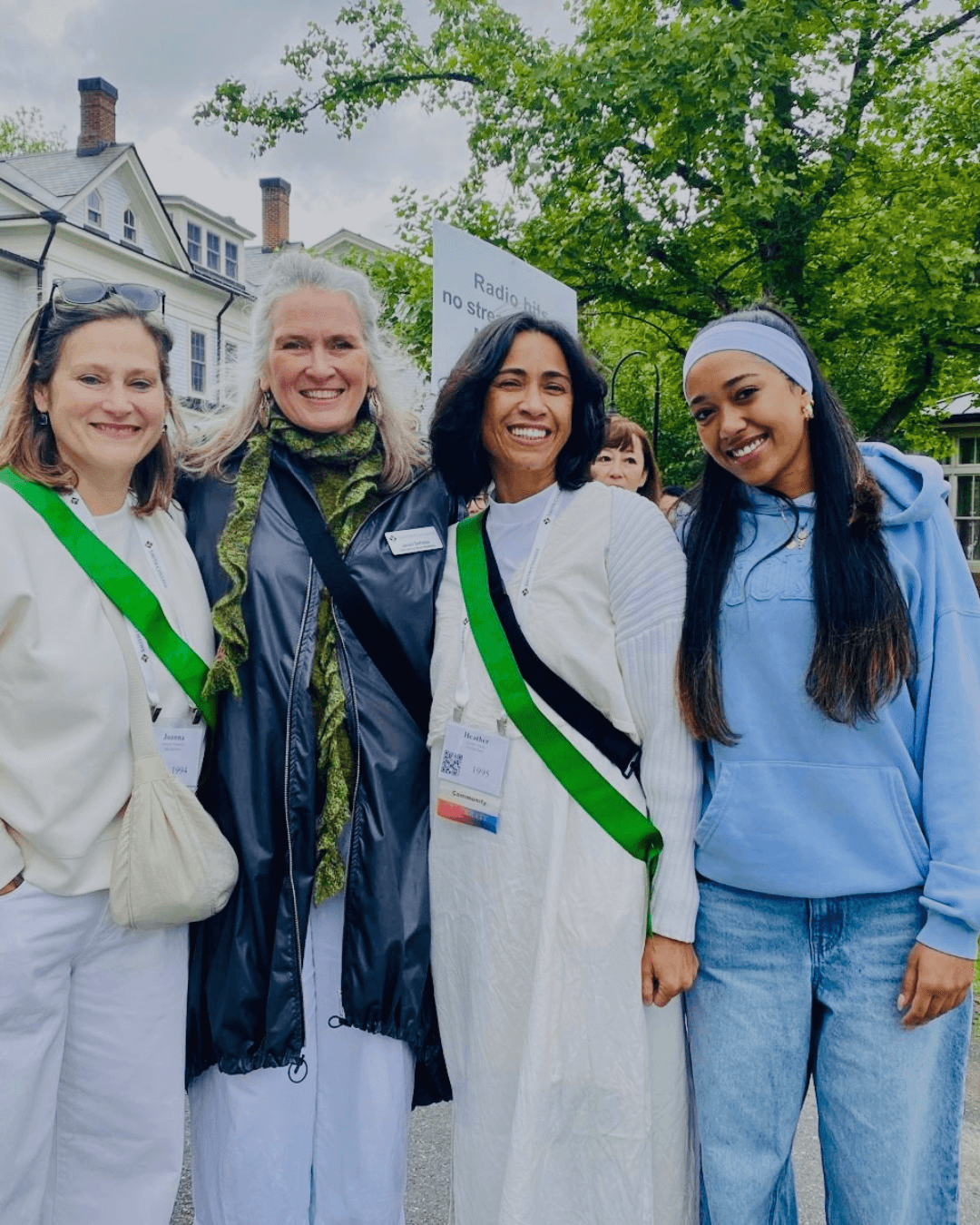 Four diverse women smile together outdoors, wearing name tags and green sashes at a reunion.