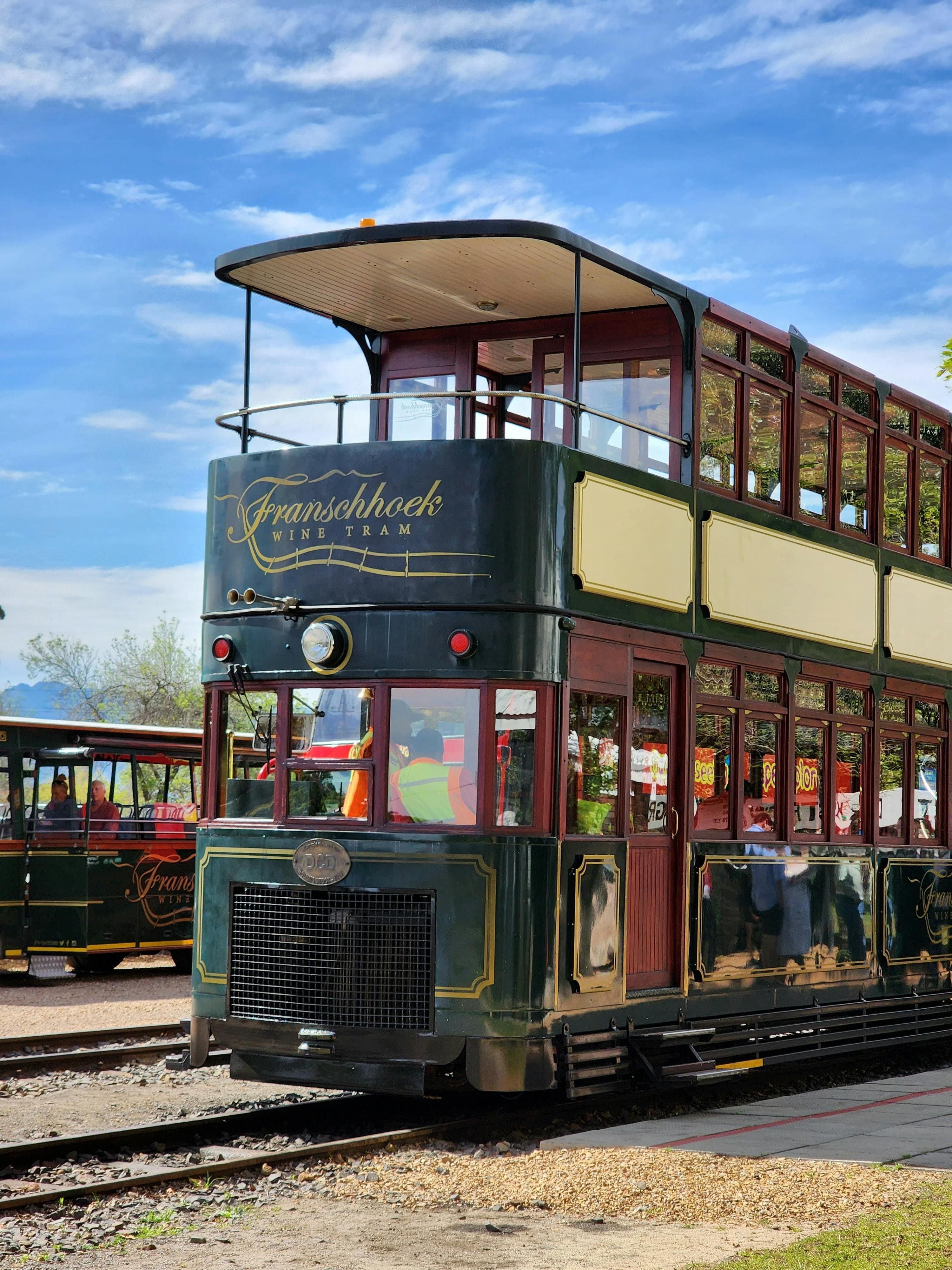Green and cream double-decker Franschhoek Wine Tram on tracks under a bright blue sky.